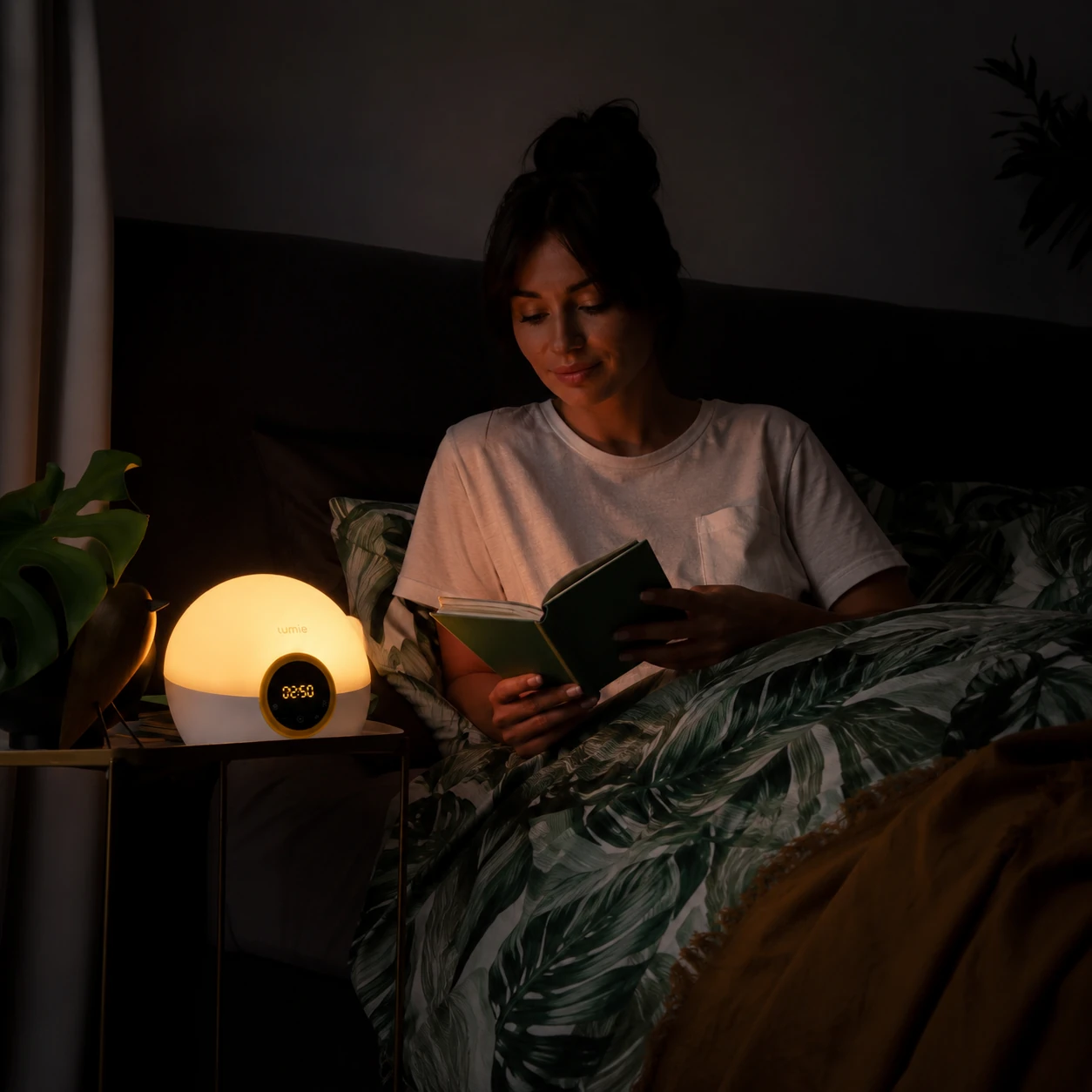 Lumie Bodyclock Rise100 sunrise alarm clock on a bedside table while a woman reads in bed under warm evening light.