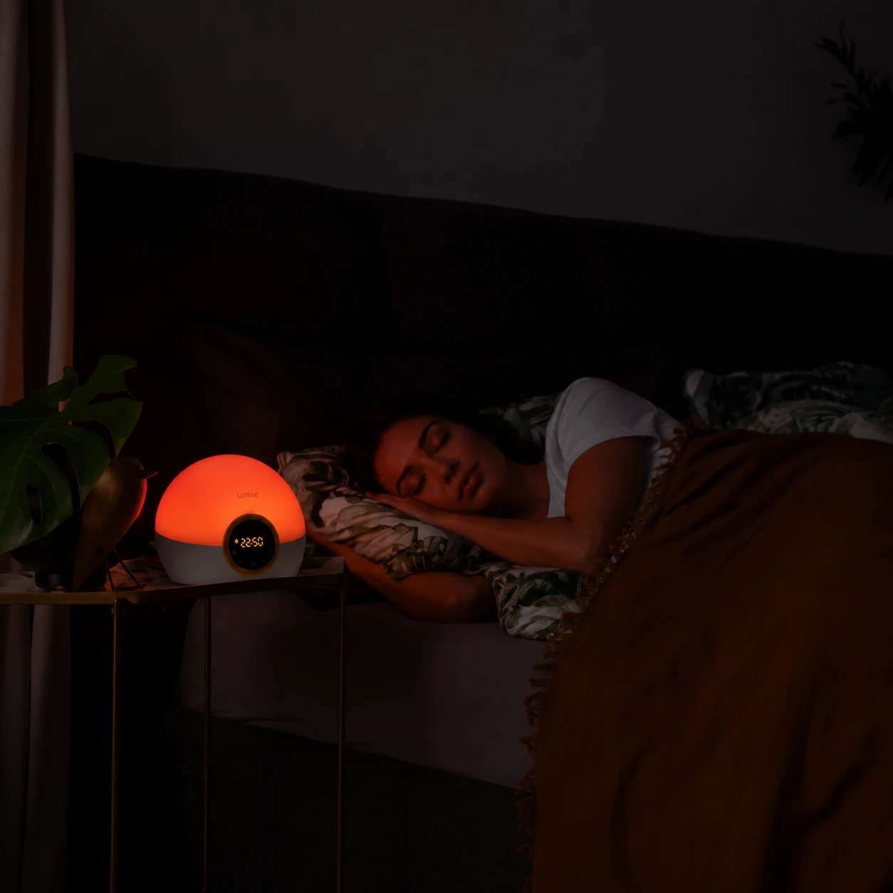 Lumie Bodyclock Rise100 alarm clock glowing red in a dark bedroom while a woman sleeps beside it.